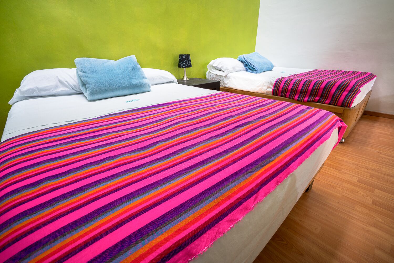 Una habitación de hotel con dos camas, una con una manta multicolor a rayas y la otra con un edredón blanco liso. Entre las camas hay una mesita de noche con una lámpara.