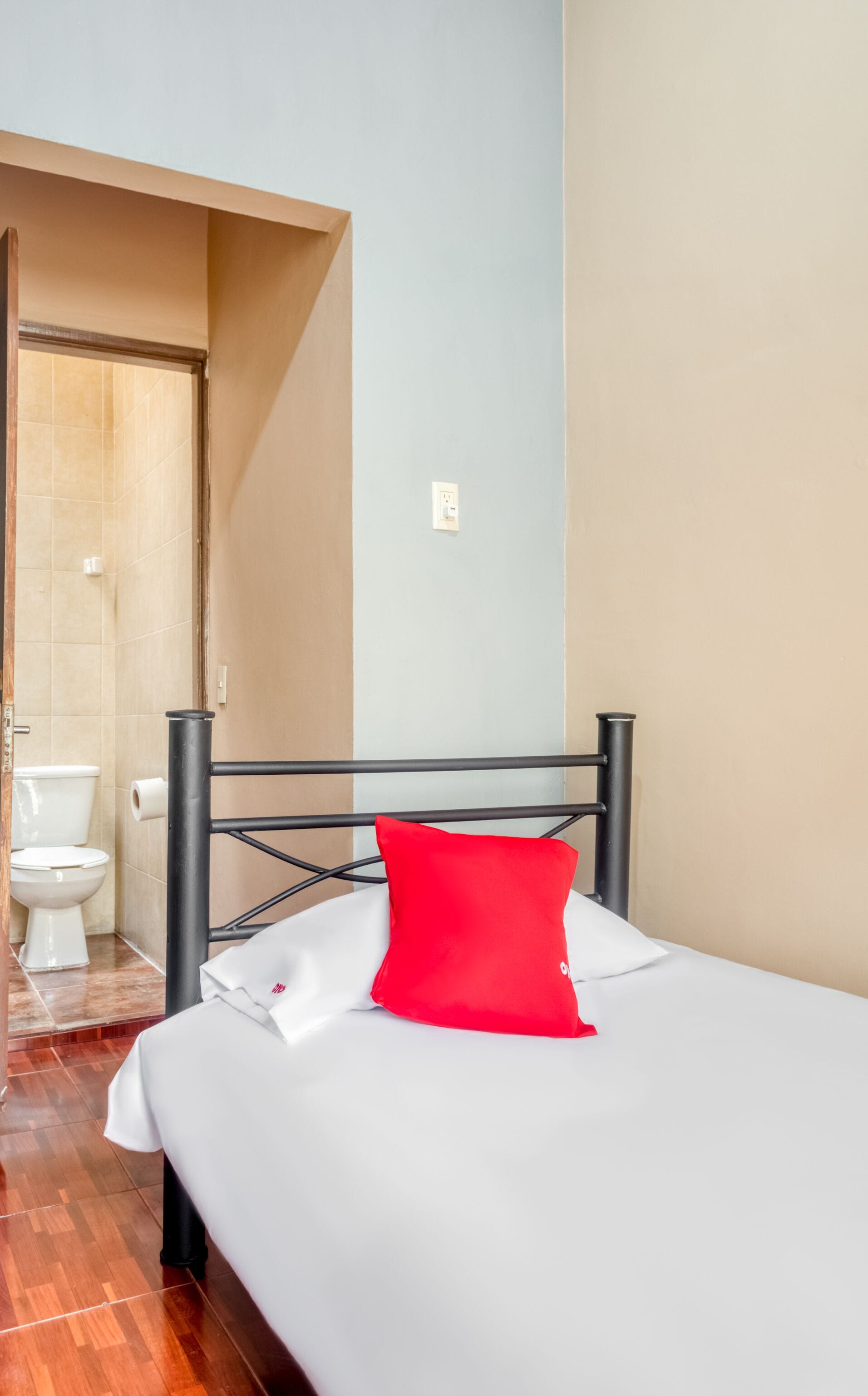 A hotel room with a bed, a red pillow, a white bedspread, a black metal headboard, a white toilet, and a shower area.