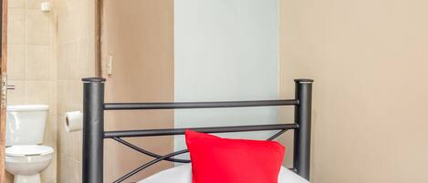 A hotel room with a bed, a red pillow, a white bedspread, a black metal headboard, a white toilet, and a shower area.