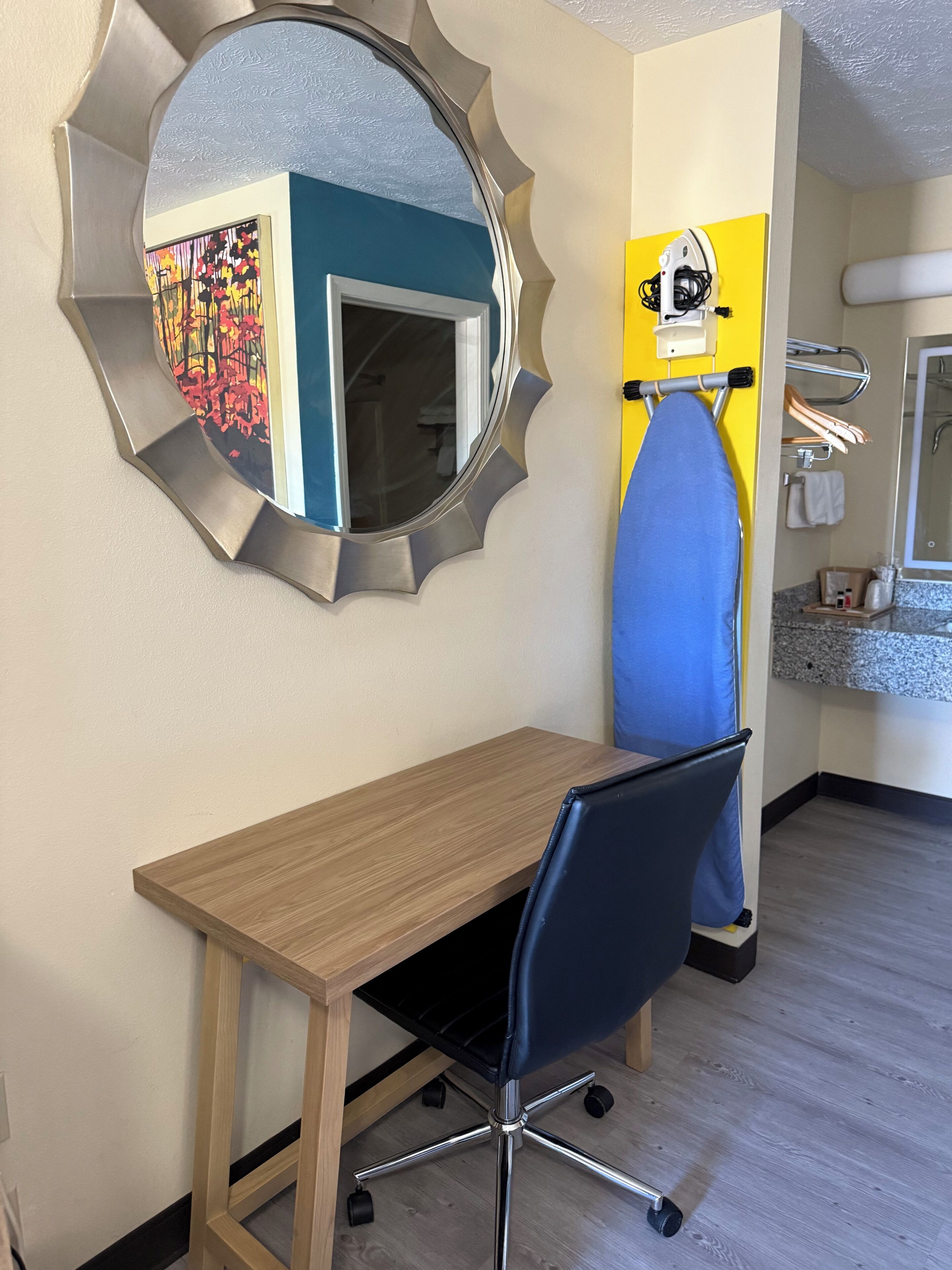 A room with a wooden desk, a blue chair, a round mirror, a yellow ironing board, and a bathroom with a sink and mirror.