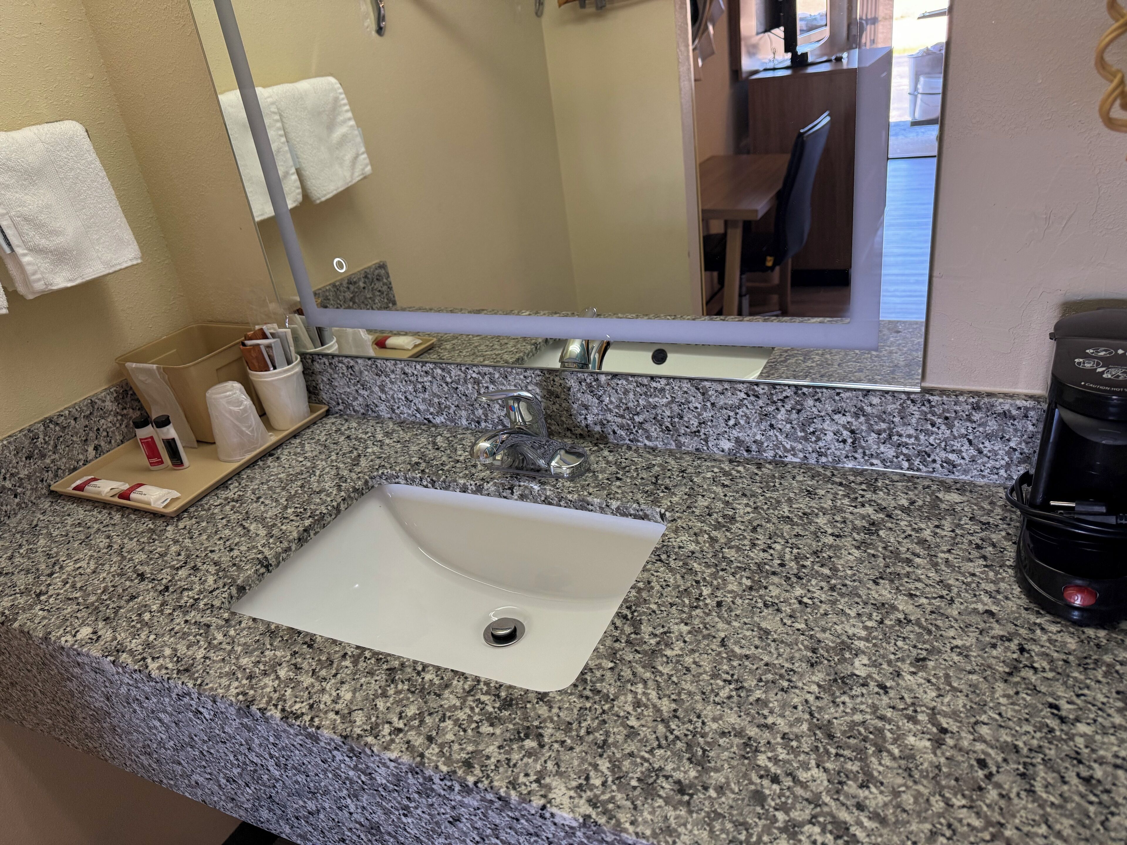 A bathroom with a granite countertop, a white sink, a mirror, and a coffee maker.