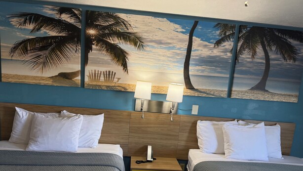 A hotel room with two beds, a nightstand, a lamp, and a wall painting of palm trees and a beach. - Deerfield Inn (Parsons)