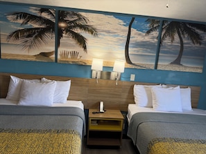 A hotel room with two beds, a nightstand, a lamp, and a wall painting of palm trees and a beach.