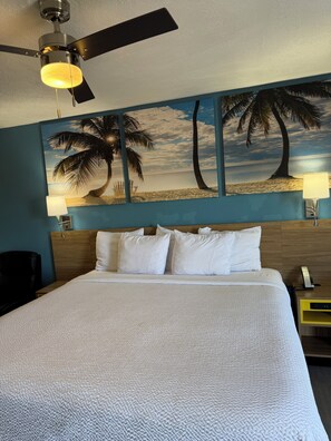 A hotel room with a bed, a ceiling fan, a painting of a beach scene, and a nightstand with a lamp.