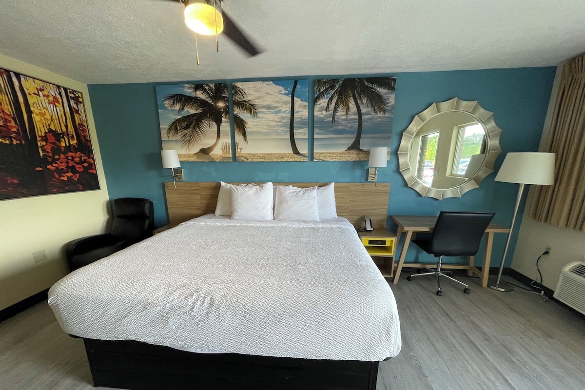 A hotel room with a large bed, a desk, a chair, a mirror, and a painting of a beach scene.