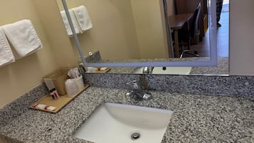 Ein Badezimmer mit einer Granit-Arbeitsplatte, einem weißen Waschbecken und einem Spiegel. Handtücher hängen an der Wand und ein kleines Regal enthält Toilettenartikel.