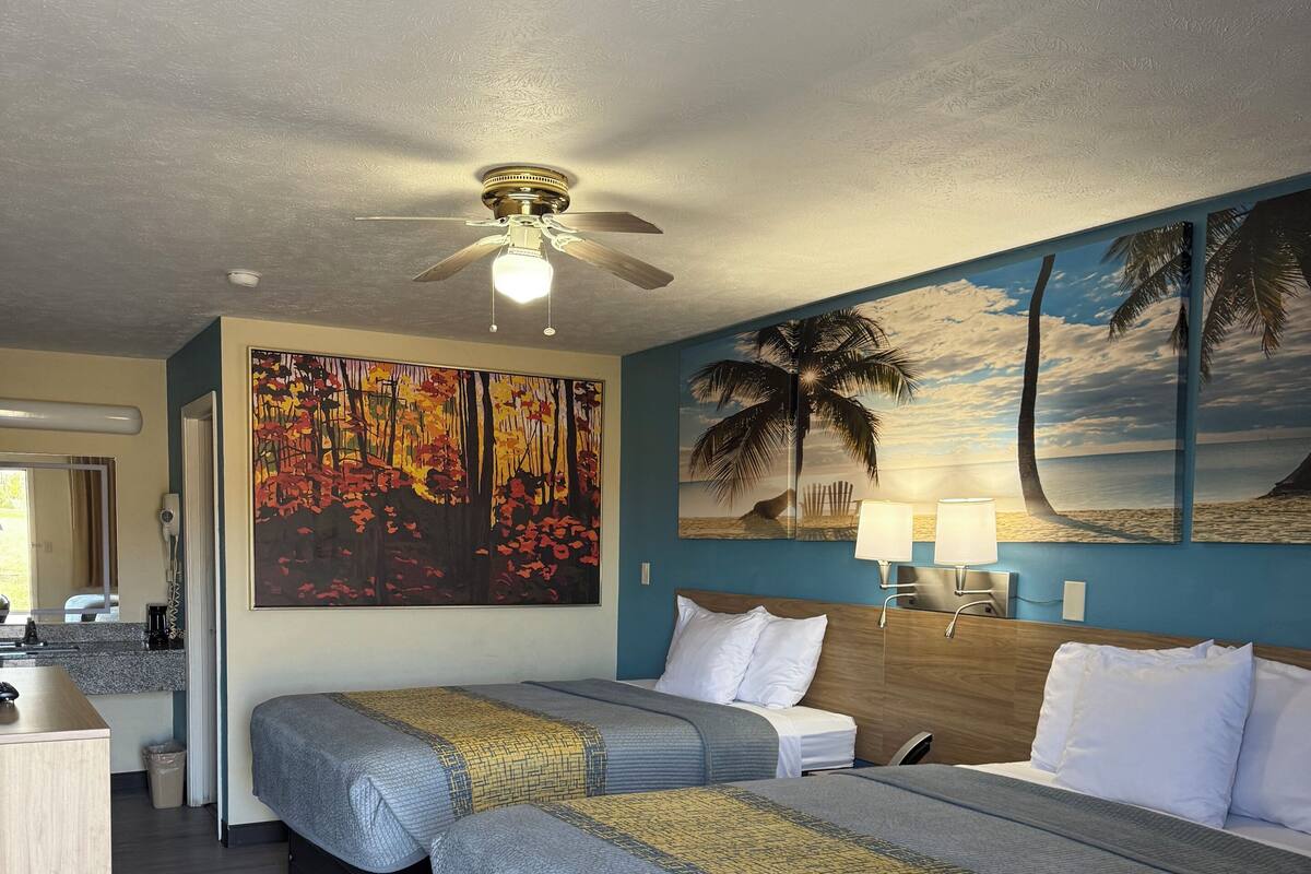 A hotel room with two beds, a ceiling fan, artwork on the walls, and a desk with a mirror.