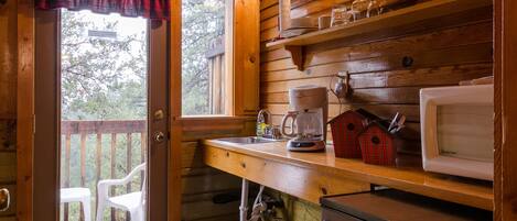 A wooden cabin kitchen with a stone tile floor, a refrigerator, a microwave, a coffee maker, and a window with a plaid curtain.