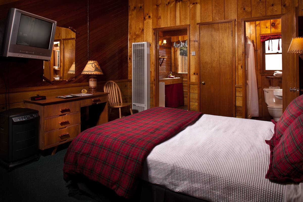 A wooden cabin room with a bed, a desk with a computer, a television, and a bathroom visible through an open door.