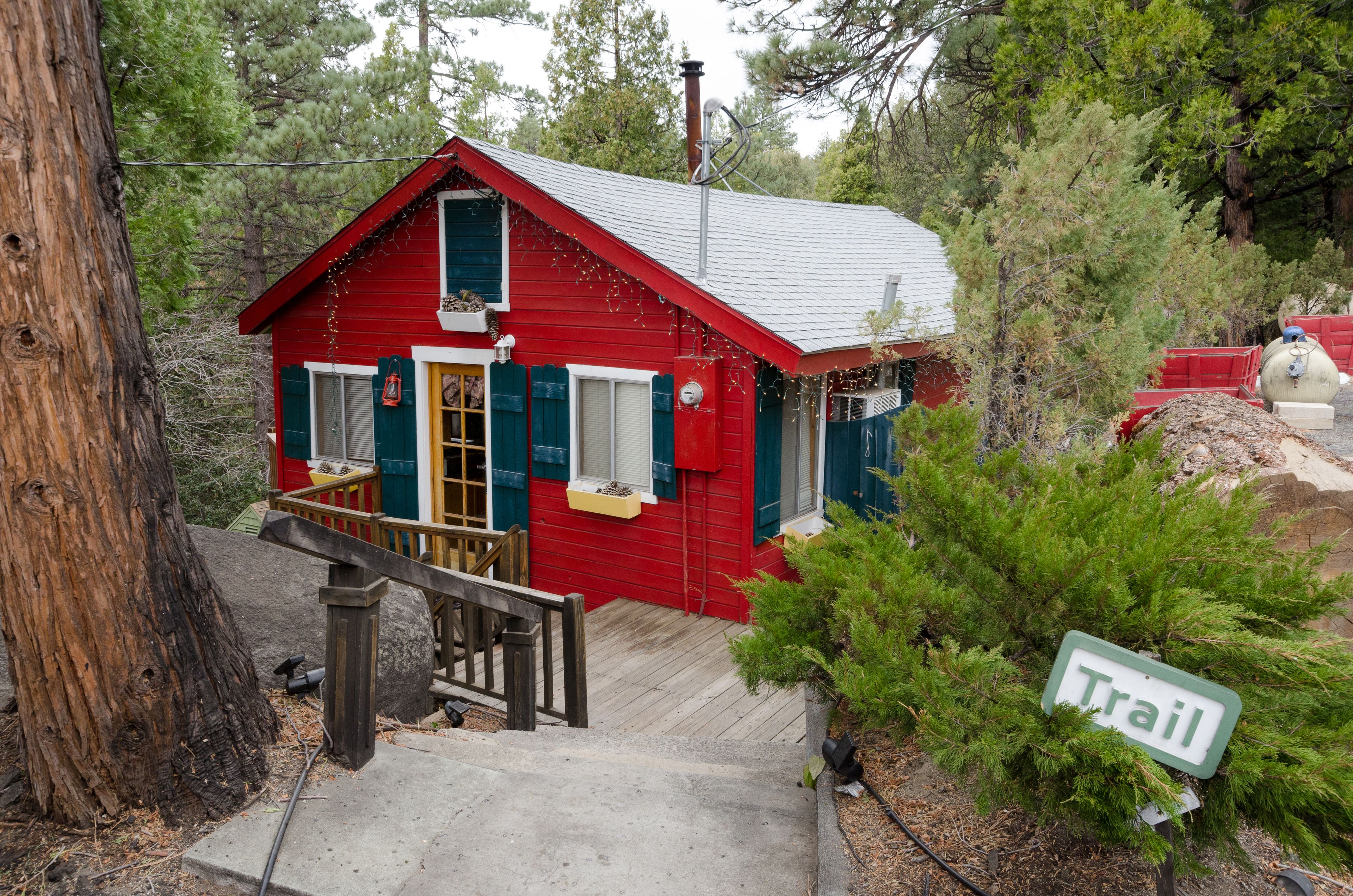 Una cabaña roja con una escalera de madera que lleva a una terraza, rodeada de árboles y un letrero que dice 'Trail'.