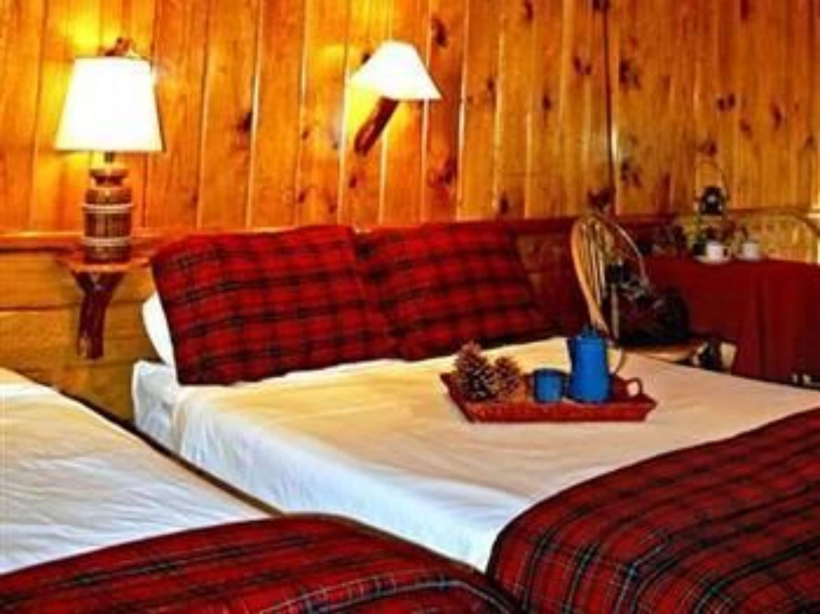 Ein Zimmer mit Holzwänden, zwei Betten, jeweils mit roter Karo-Bettwäsche, einem kleinen Tisch mit Lampe und einer Ablage mit Gegenständen.