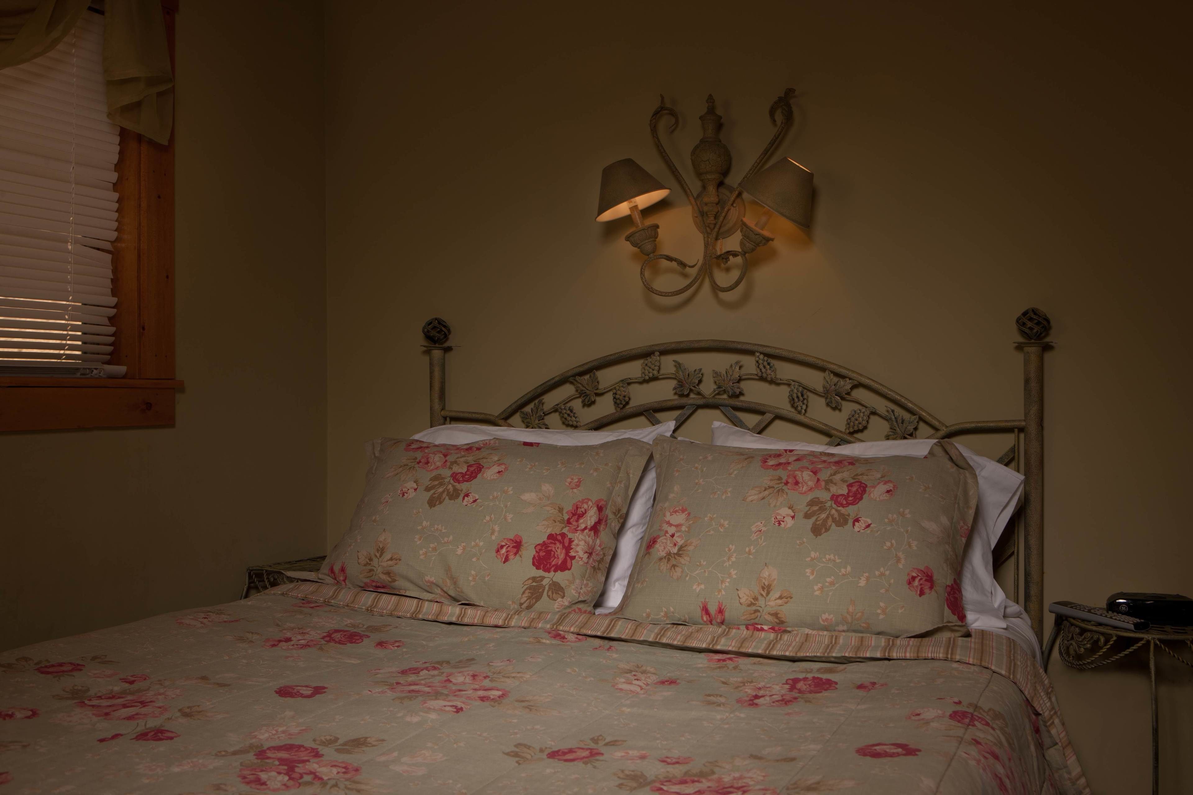 Una cama con ropa de cama floral, un aplique de pared y una ventana con persianas.