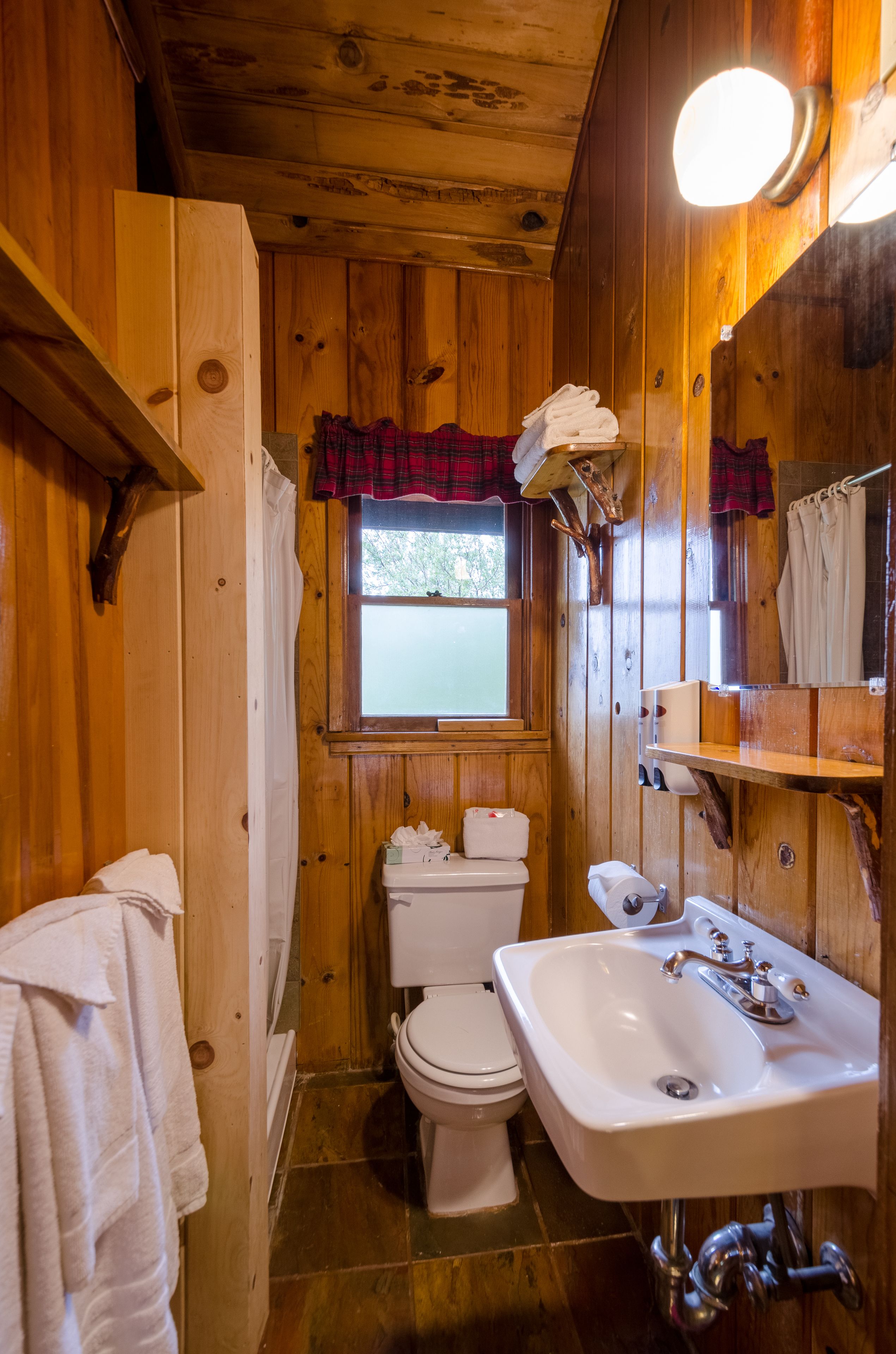 Baño de cabaña de madera con inodoro, lavamanos y ventana con cortina escocesa.