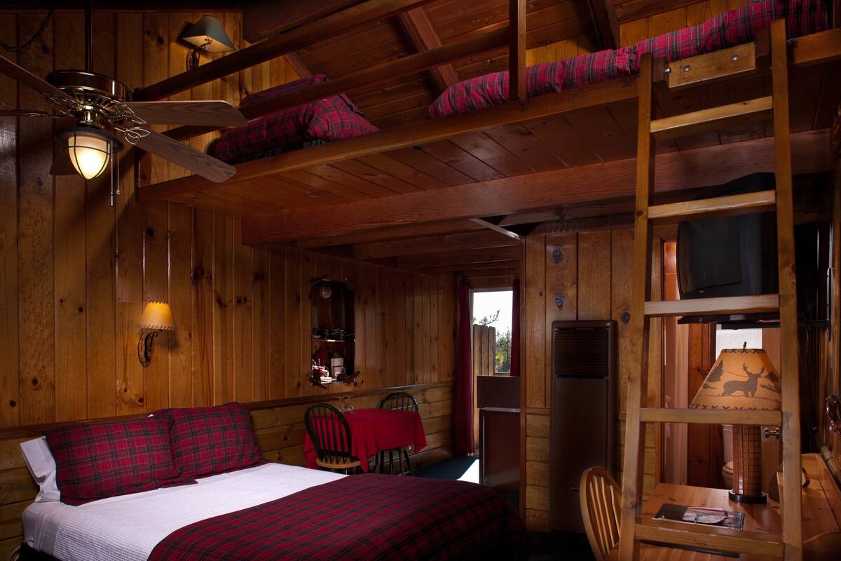 A cabin-style room with a bed, a desk, and a chair. There is a ceiling fan, a window, and a door.