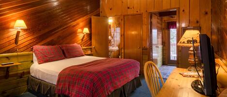 A cozy wooden cabin bedroom with a bed, two bedside lamps, a wooden desk with a computer, and a door leading to another room.