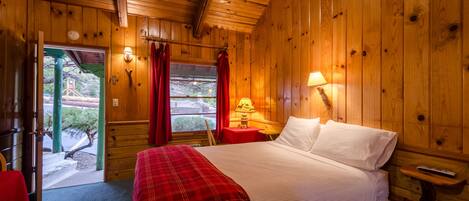 A wooden room with a bed, a red plaid bedspread, a nightstand with a lamp, and a door leading outside.