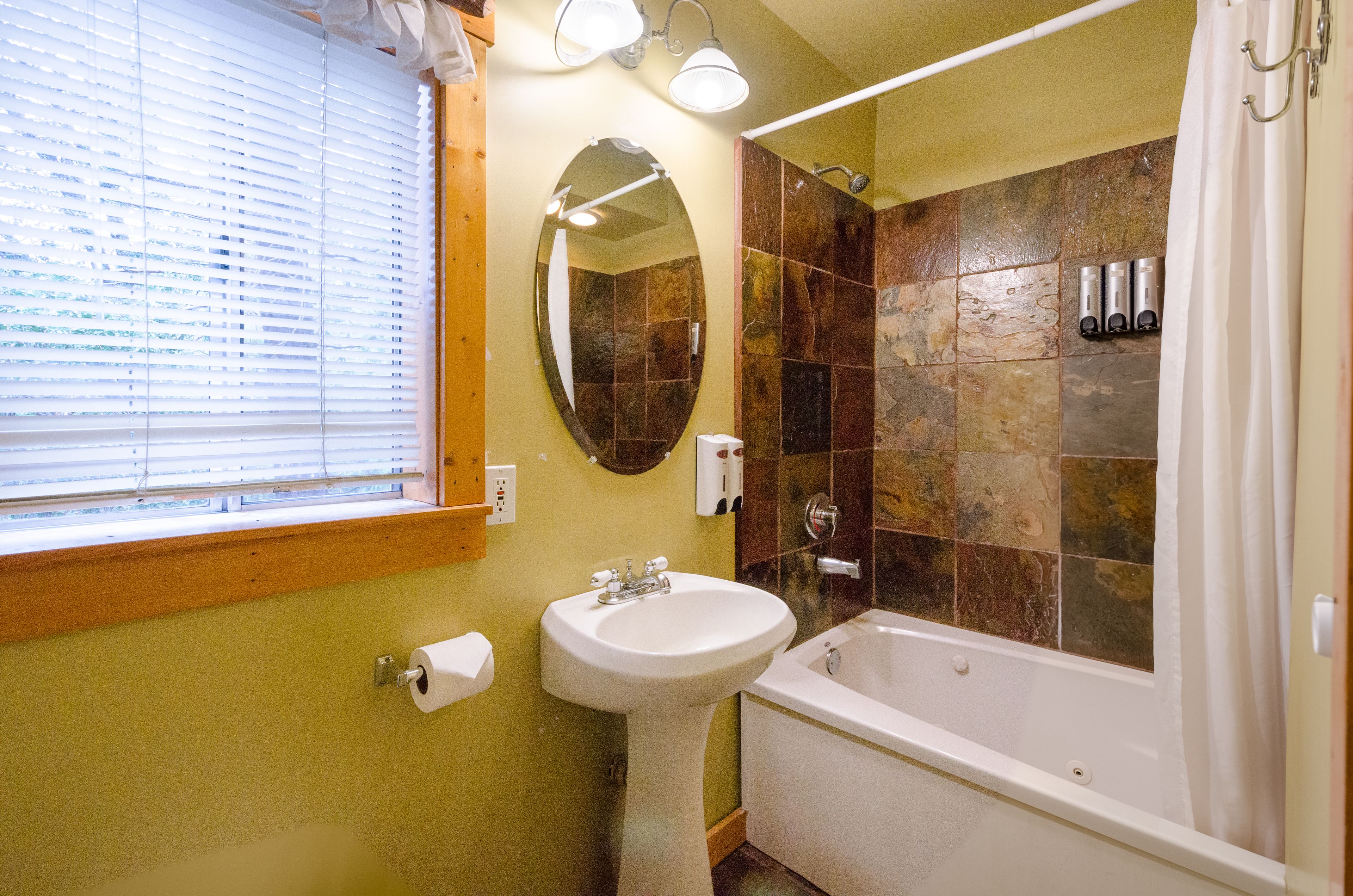 A bathroom with a pedestal sink, a bathtub, a shower with stone tiles, a window with blinds, and a mirror.