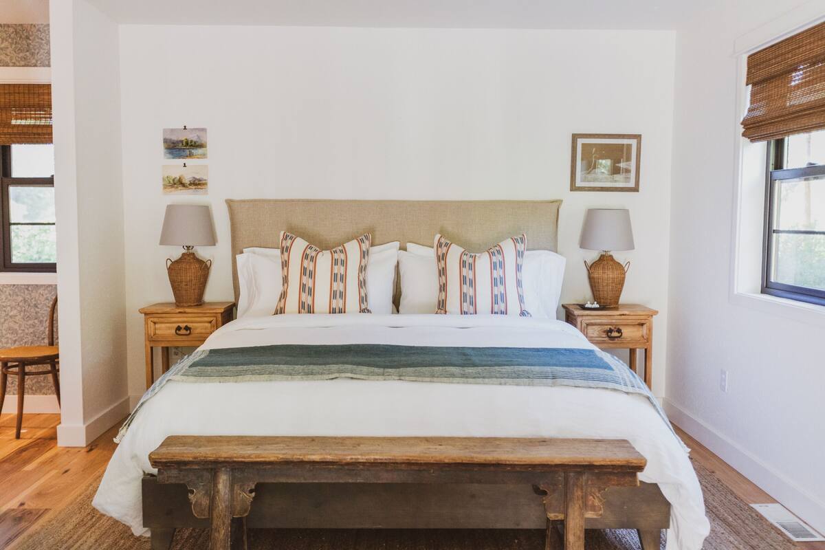Un dormitorio con una cama grande, dos mesitas de noche, un banco de madera y dos lámparas de mimbre.