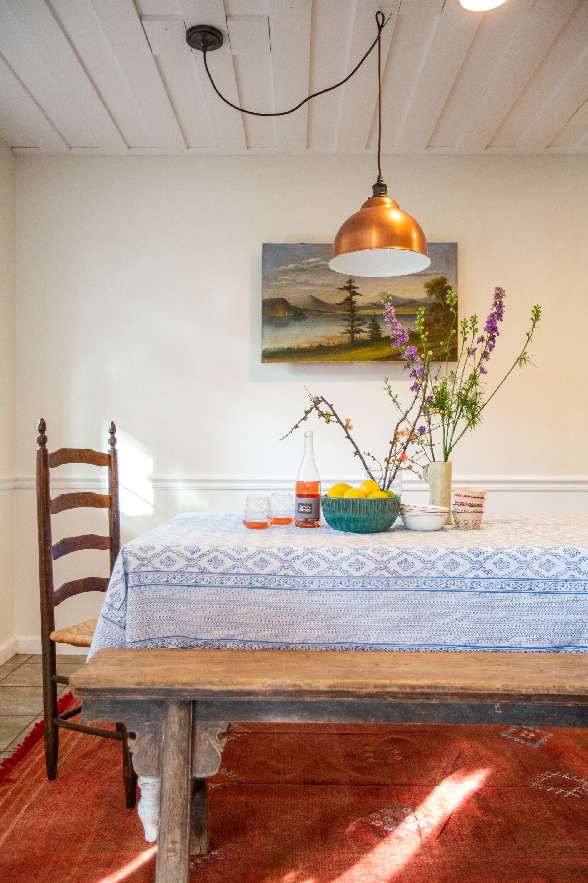 Un comedor con una mesa de madera, una silla, un jarrón con flores, una botella y un cuadro en la pared.