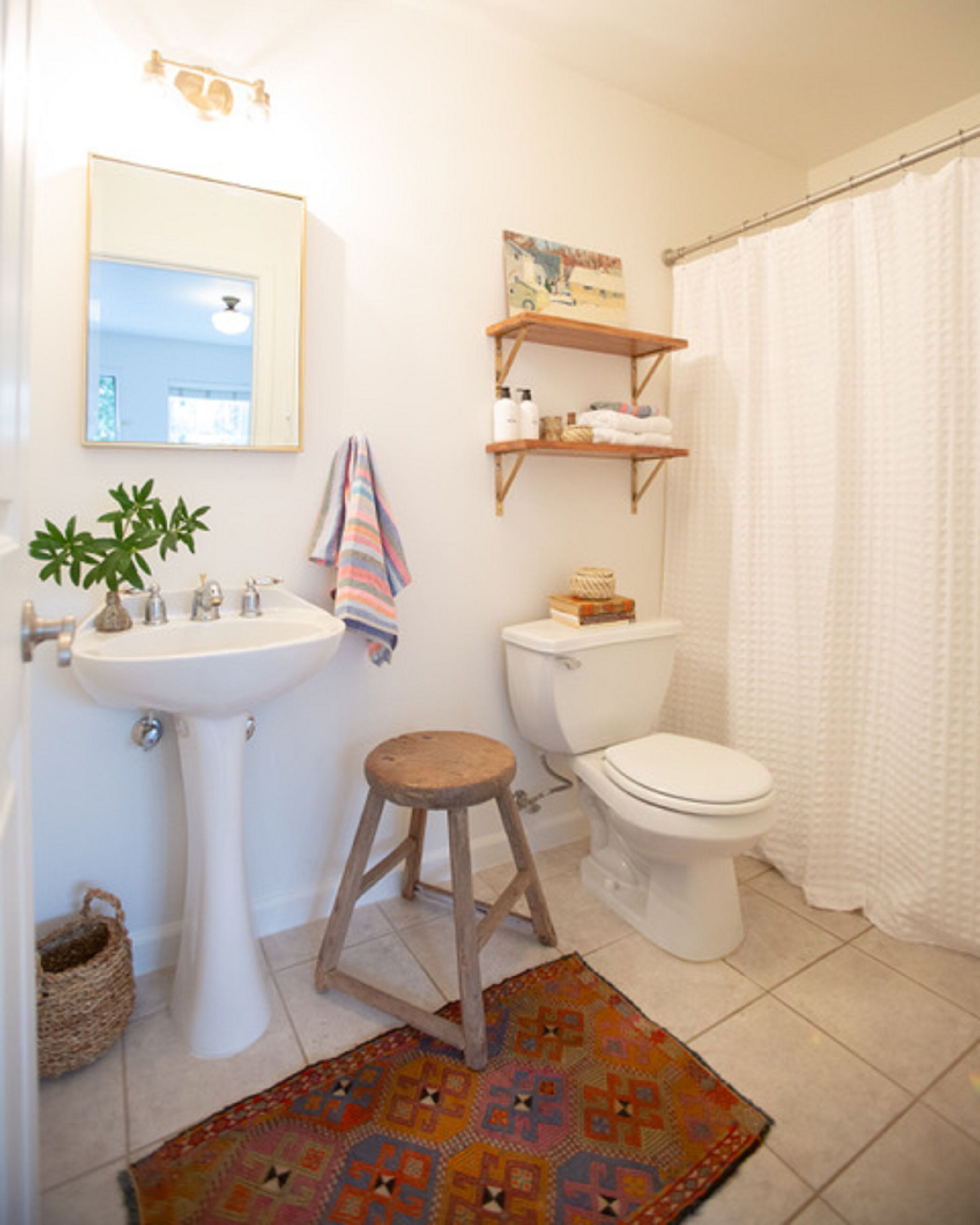 Une salle de bain avec un lavabo sur colonne, des toilettes et un rideau de douche. On y trouve un petit tabouret et un tapis à motifs sur le sol.