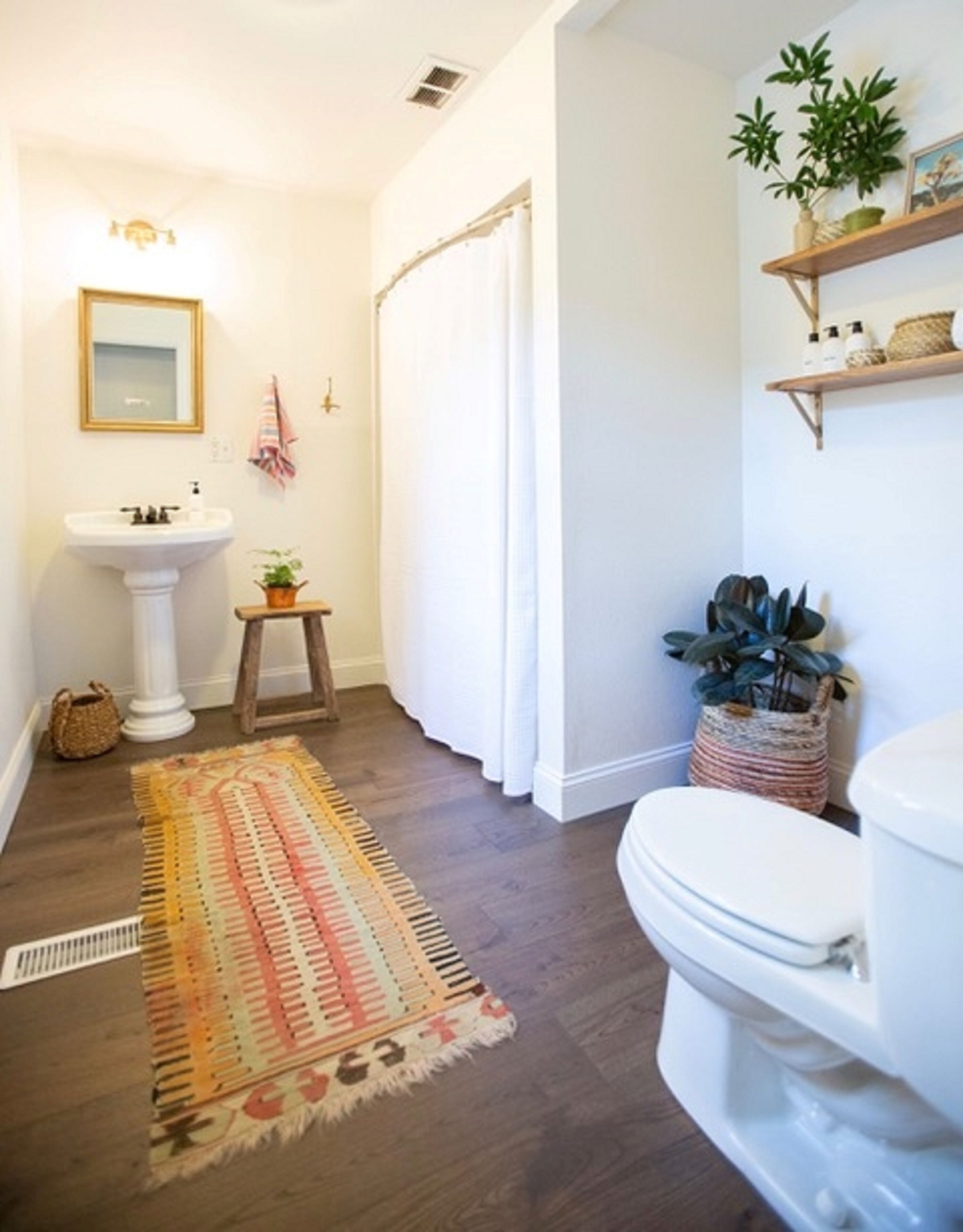 Un baño con lavamanos pedestal, inodoro, cortina de ducha, alfombra y estantes con plantas.