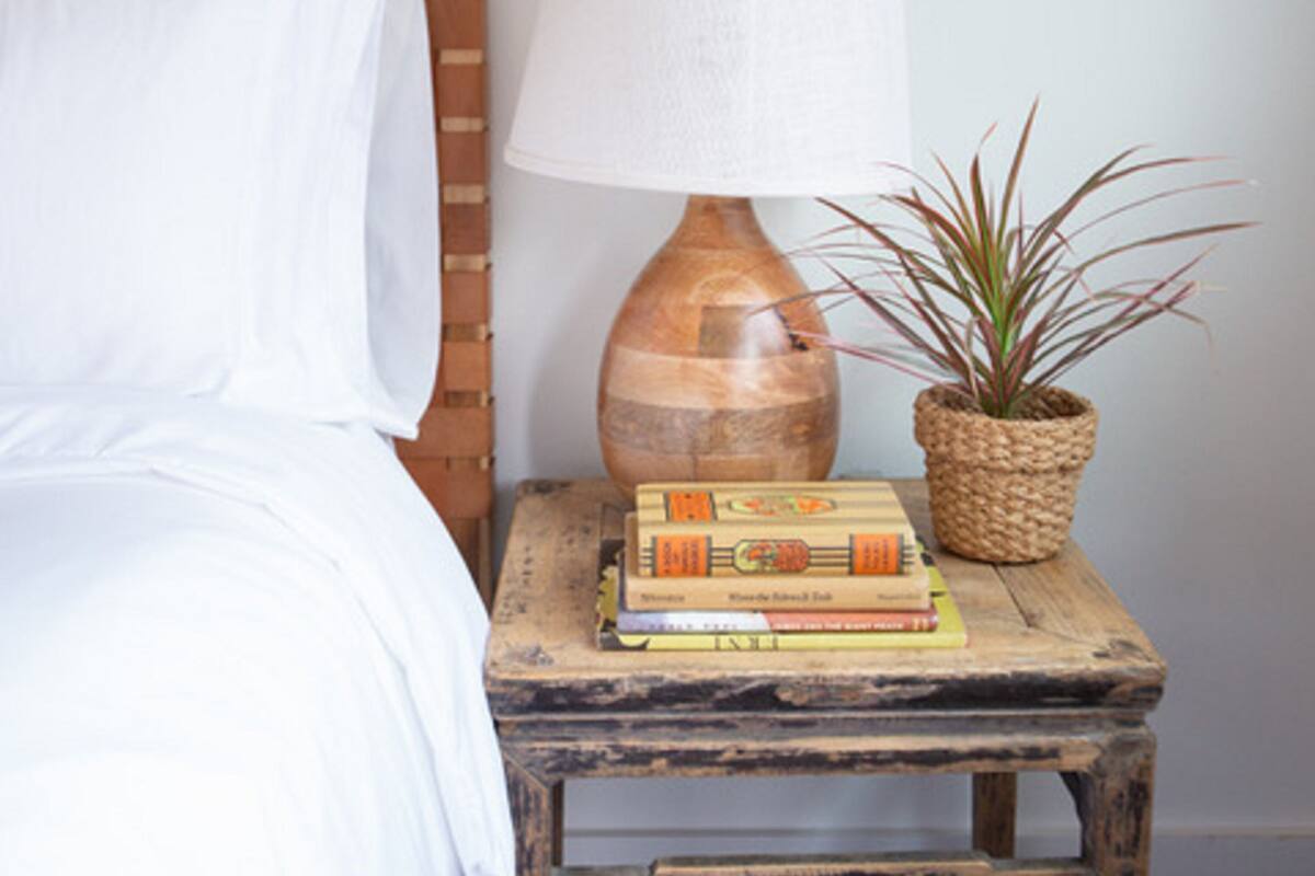 Una cama con ropa de cama blanca, una mesita de noche de madera con una lámpara y libros, y una planta en maceta.