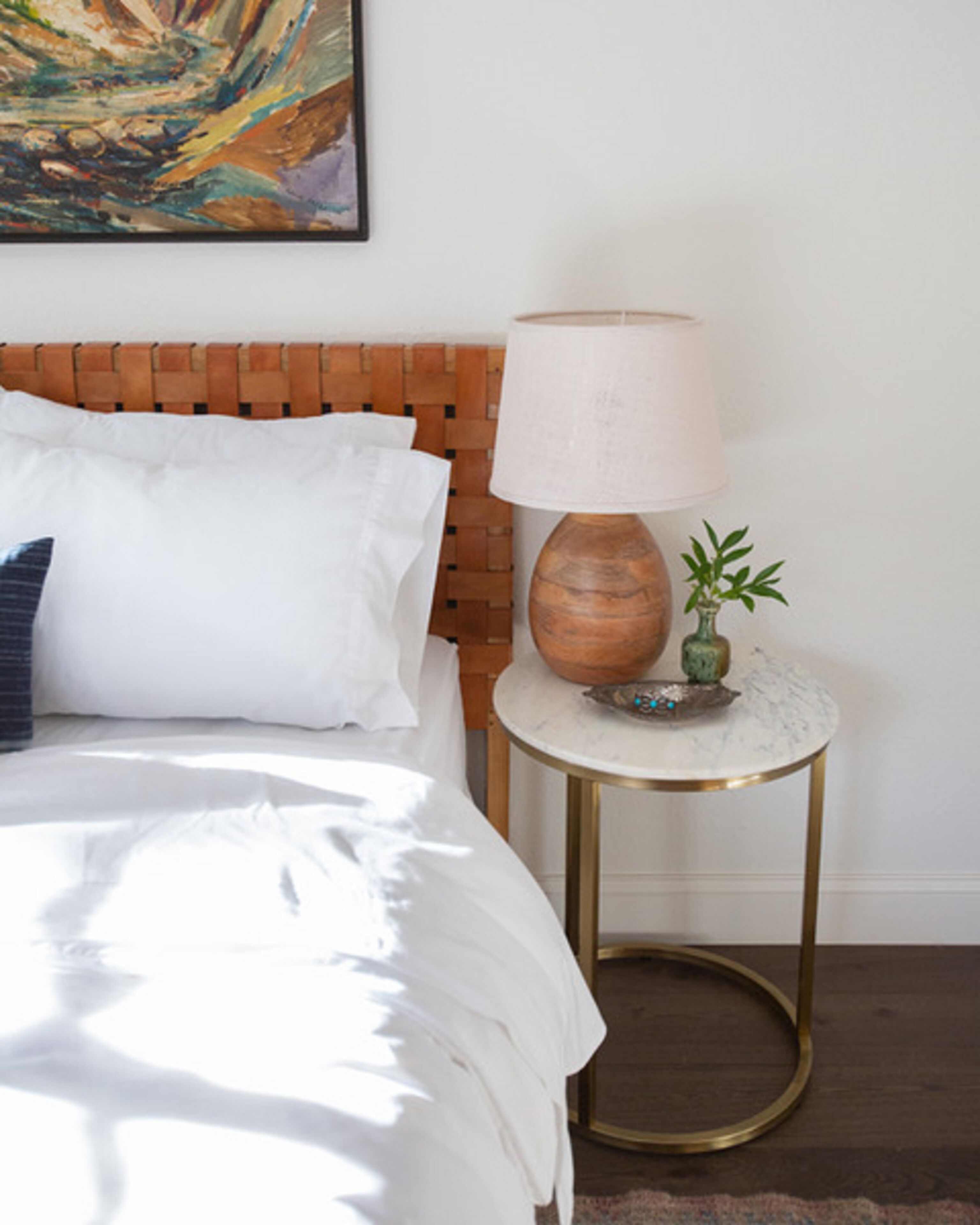 A neatly made bed with white bedding and a blue pillow, a wooden headboard, a small round table with a lamp, and a small potted plant.
