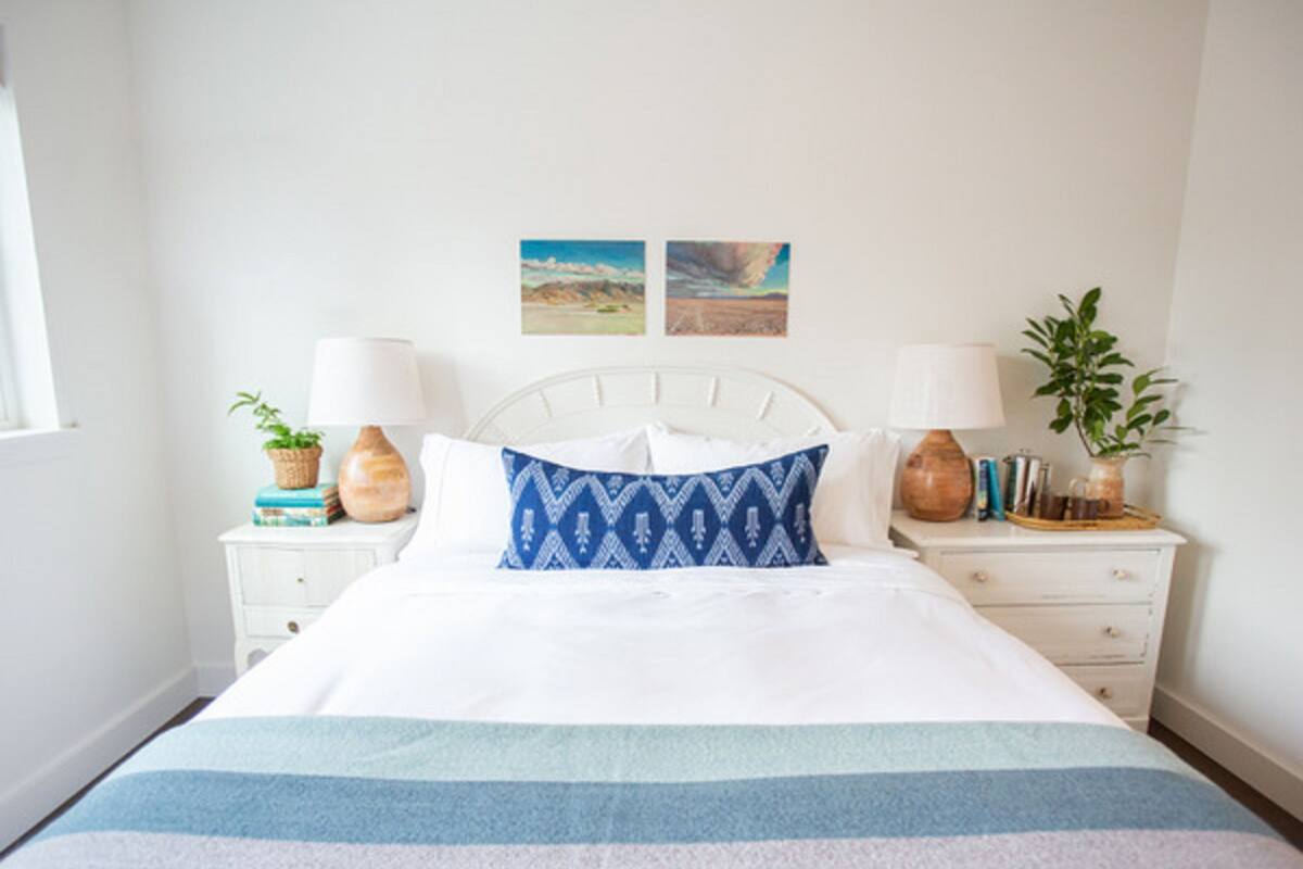 Una cama bien hecha con una colcha a rayas, dos mesitas de noche con lámparas y objetos decorativos, y dos cuadros en la pared.
