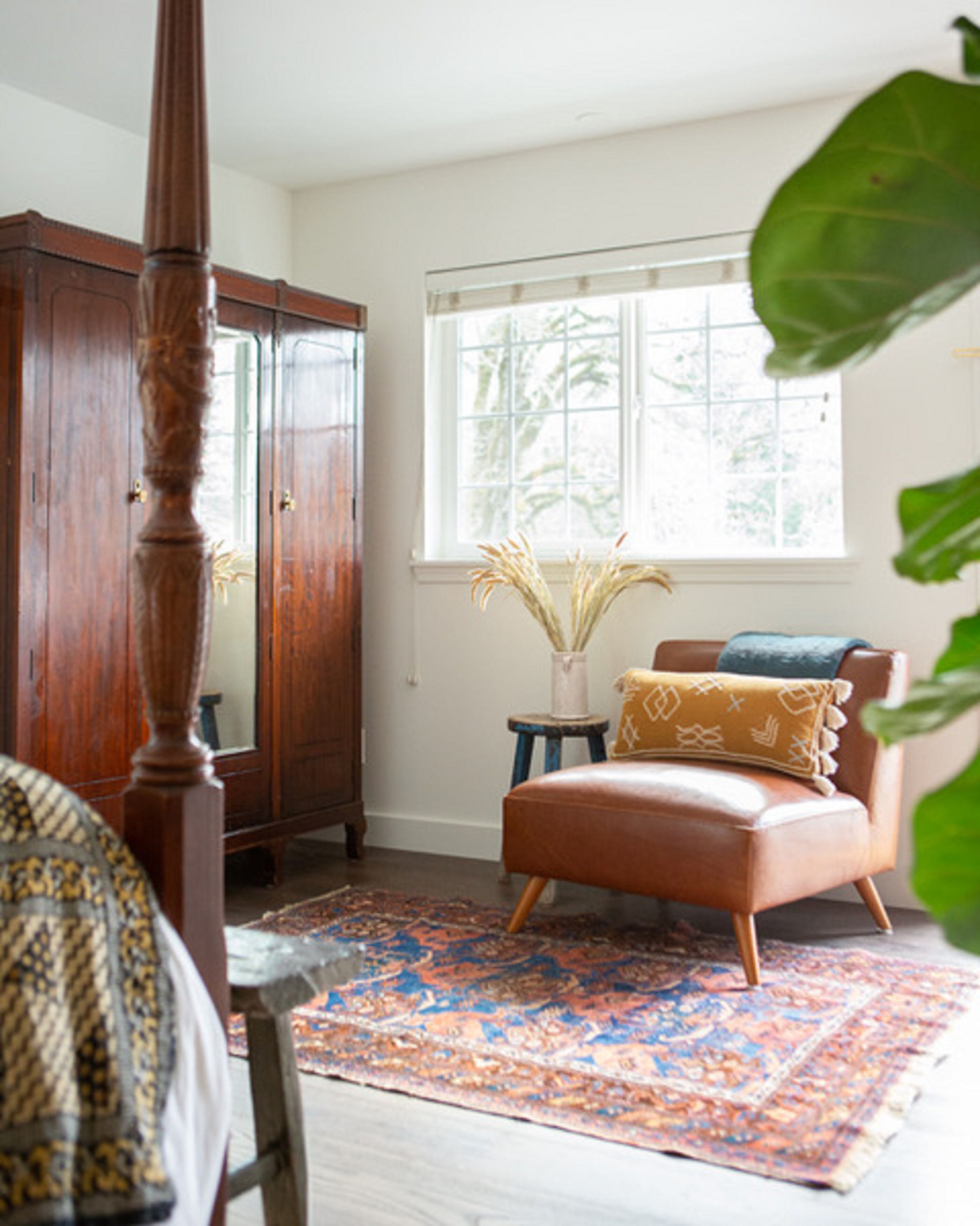 A room with a wooden wardrobe, a brown leather armchair, a patterned rug, and a window with a view of trees.