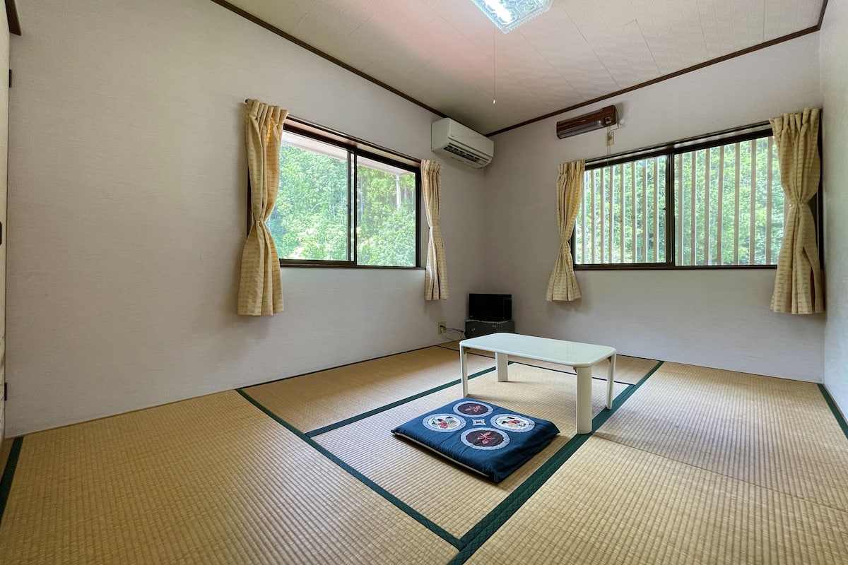 Een kamer met tatami vloermatten, een witte tafel en twee ramen met gordijnen.
