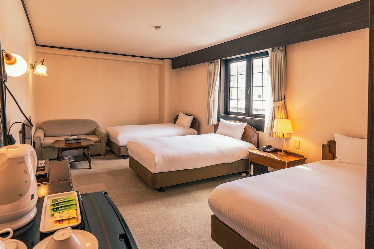 A hotel room with two beds, a sofa, a desk with a lamp, and a window with curtains.