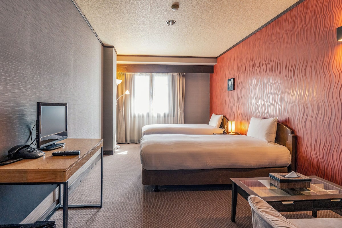 A hotel room with a bed, a desk with a computer and phone, a bedside table with a lamp, and a view of the outdoors.
