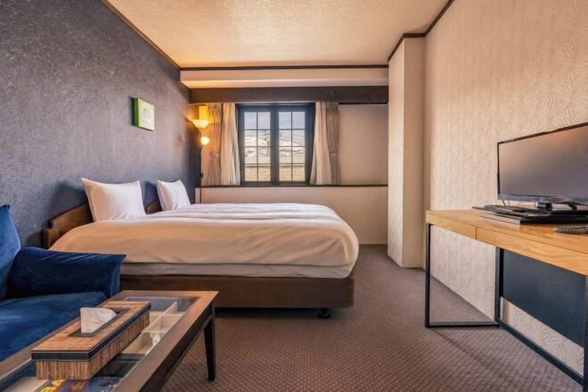 A hotel room with a bed, a TV, a desk, and a window with curtains.