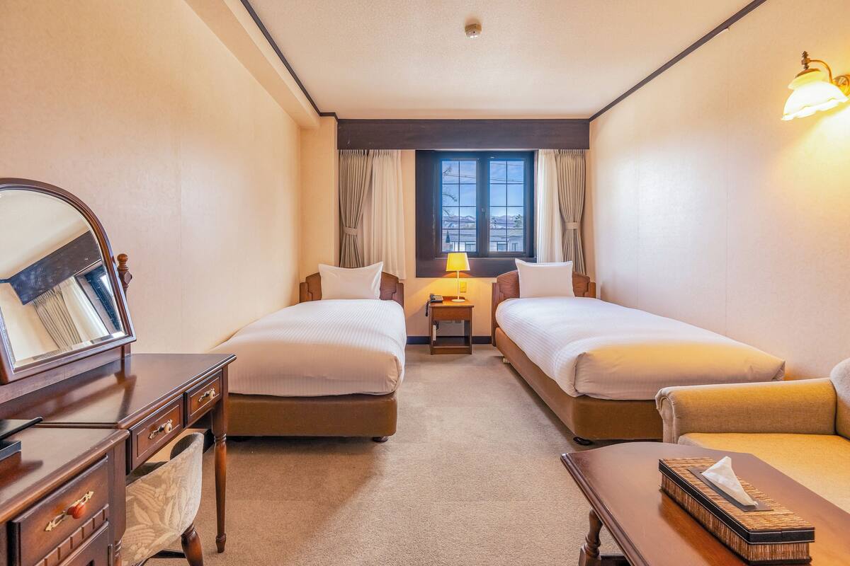 A hotel room with two beds, a desk with a mirror, a lamp, and a view of the outdoors through the window.