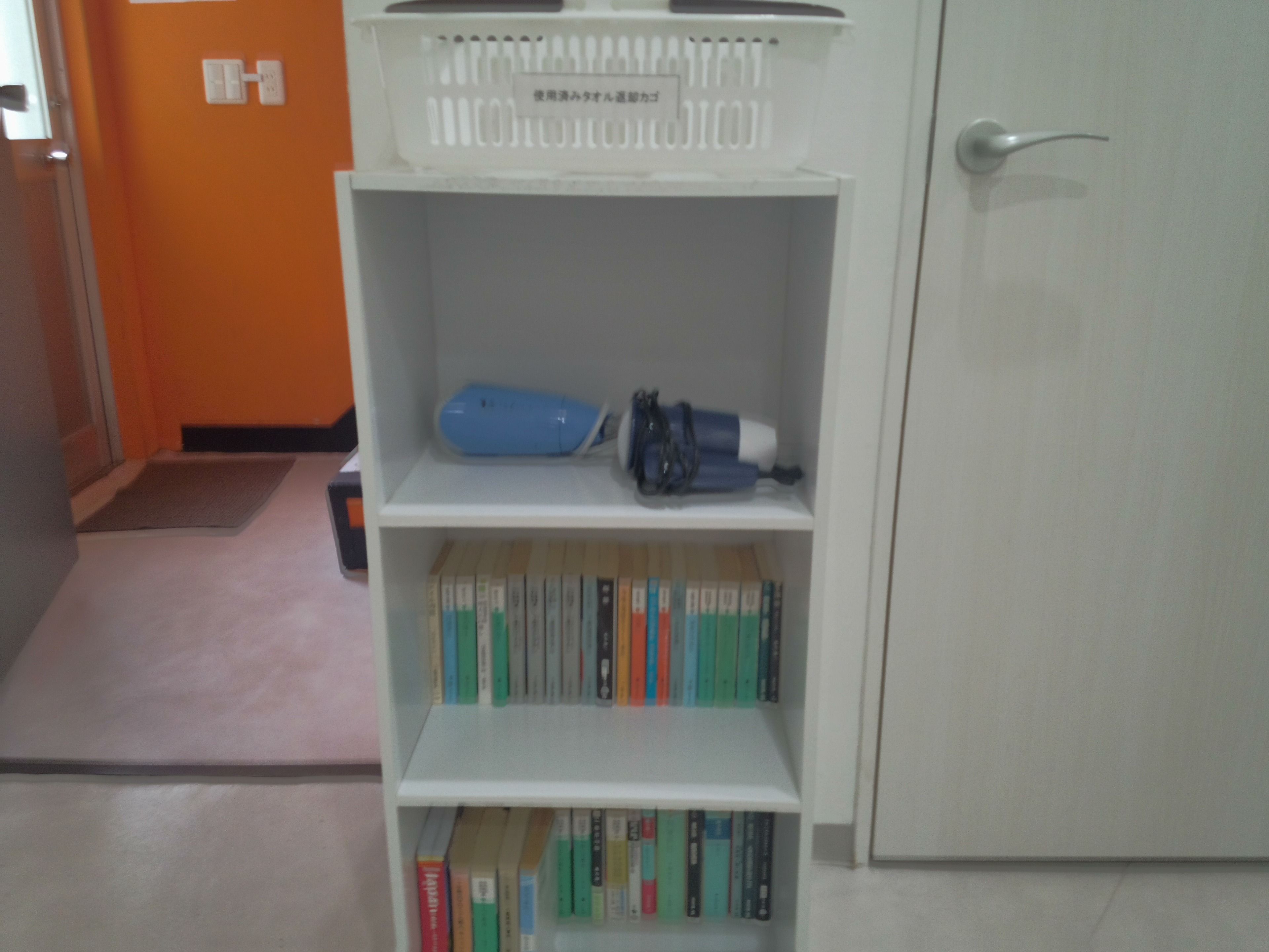 Una estantería blanca con libros y un objeto azul en el estante superior, una puerta blanca a la derecha y una pared naranja al fondo.