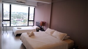A modern hotel room with two beds, a large window offering a city view, an air conditioning unit, and a small table with a chair. - Marina Travellers Suite SKY Karamunsing (Kota Kinabalu)