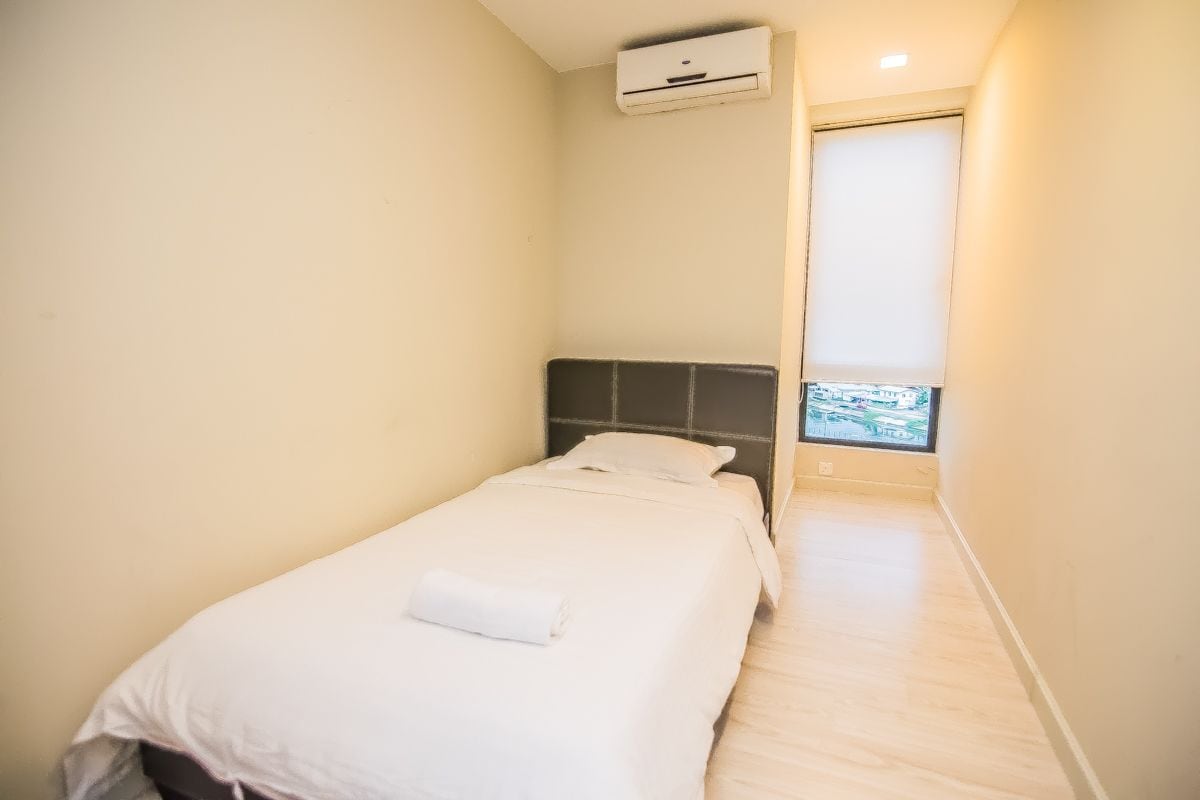 A small, single-bed room with a plain white bedspread, a dark headboard, and a window with a view of buildings.