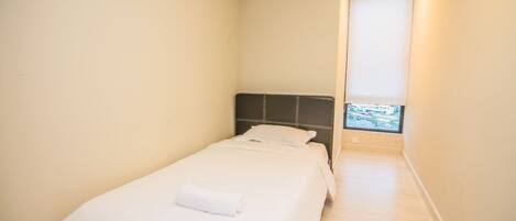 A small, single-bed room with a plain white bedspread, a dark headboard, and a window with a view of buildings.