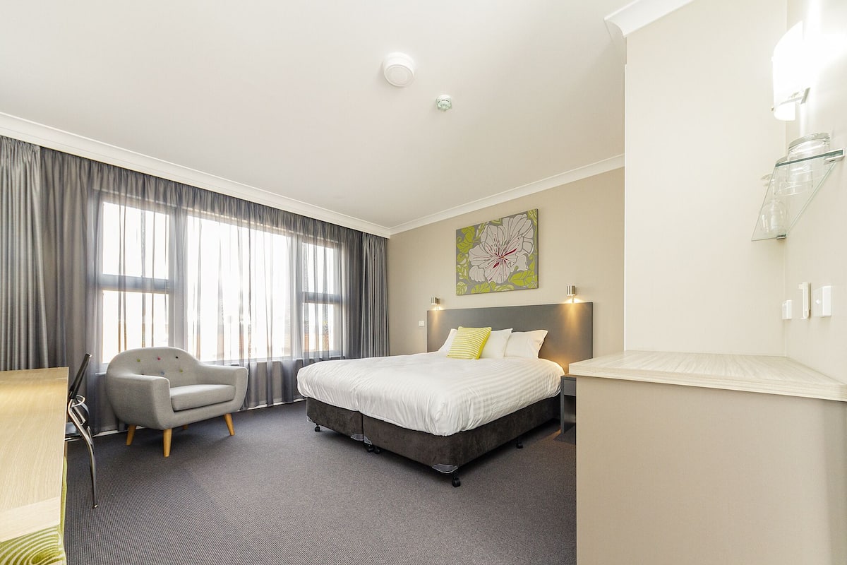 A modern hotel room with a large bed, a grey armchair, a desk, and a large window with curtains.