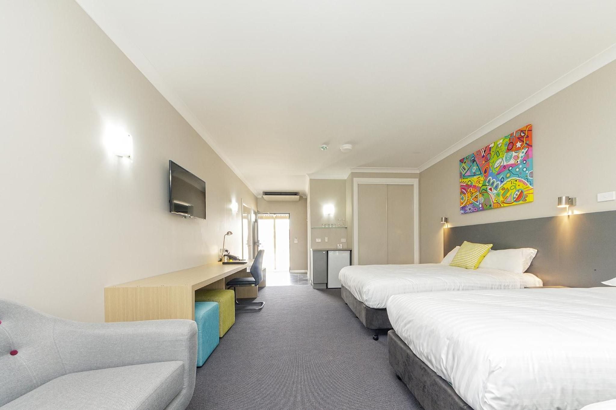 Ein Hotelzimmer mit zwei Betten, einem Schreibtisch, einem Fernseher und einem farbenfrohen Kunstwerk an der Wand.