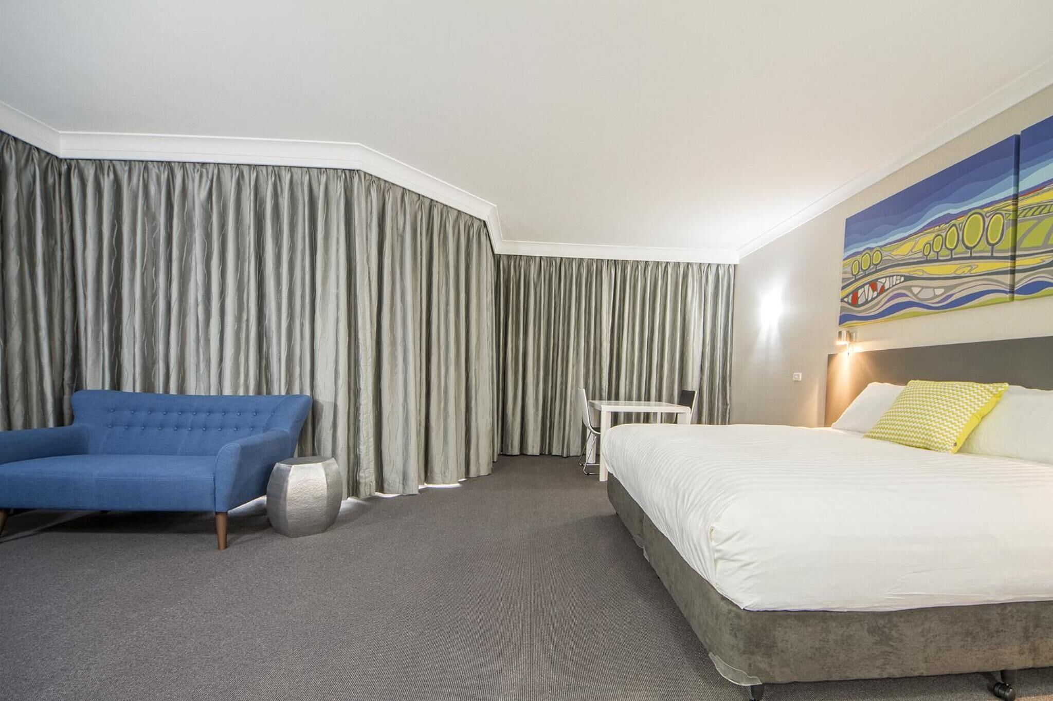 A hotel room with a large bed, a blue armchair, a small table, and a painting on the wall.