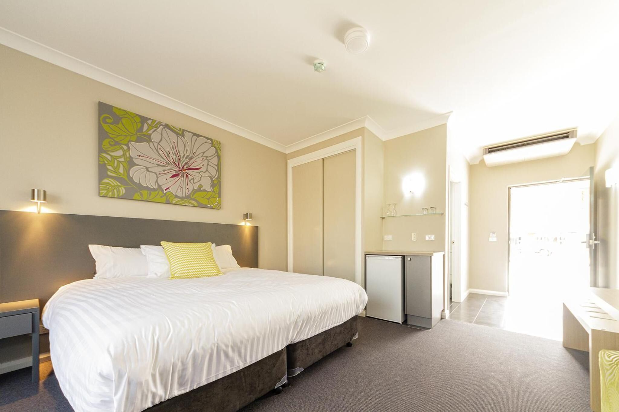 A hotel room with a large bed, a bedside table, a small desk, a mirror, and a door leading to the bathroom.