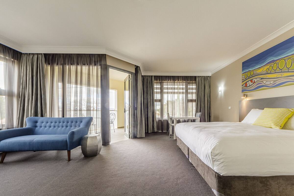 A hotel room with a large bed, a blue armchair, a small table, and a painting on the wall.
