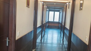 A long, narrow hallway with dark wooden doors and walls, leading to a bright room at the end. - Hotel Taj Heights (Agra)