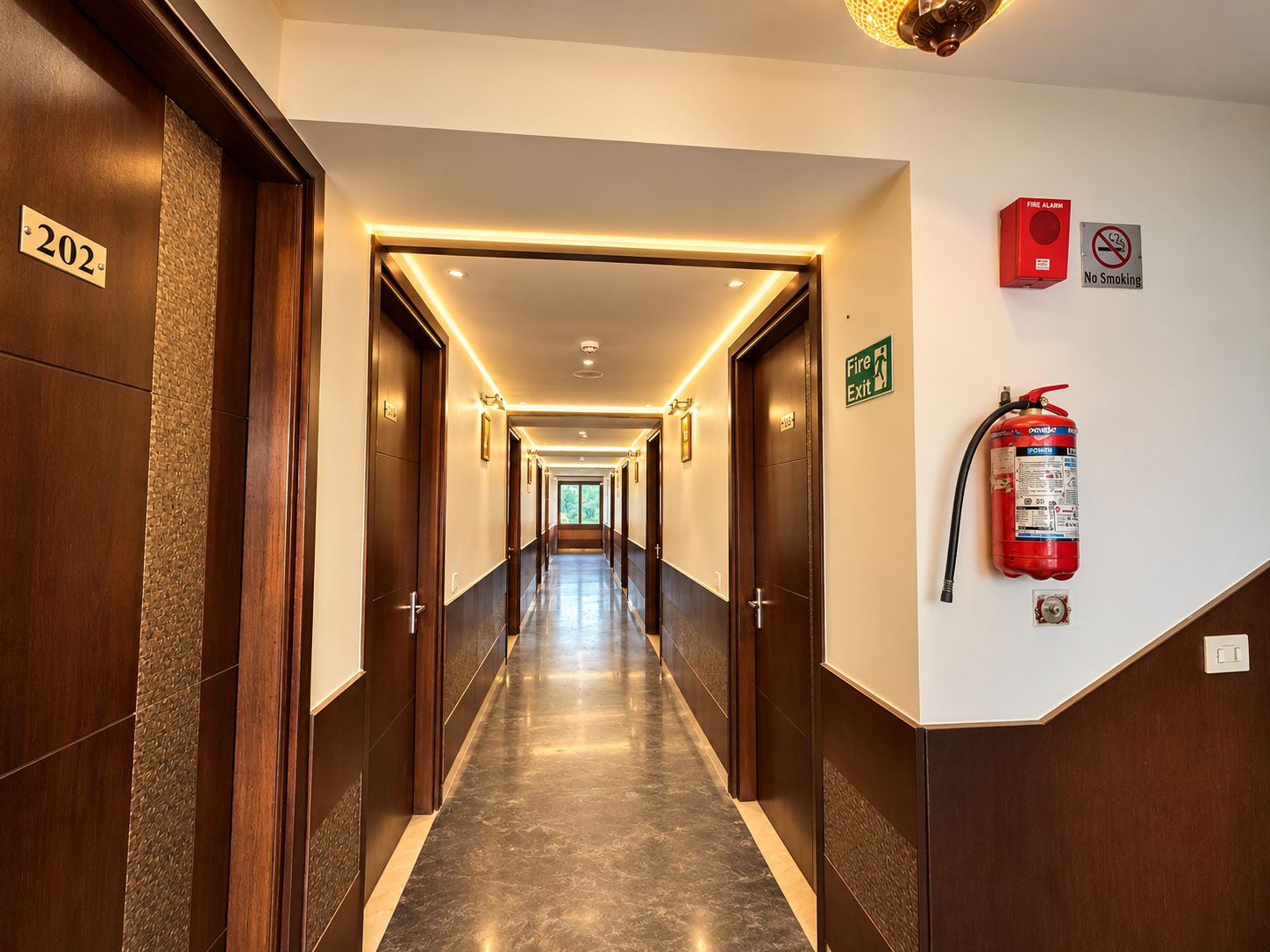 A hotel corridor with doors numbered 202, fire exit signs, a fire alarm, and a fire extinguisher.