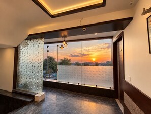 A modern interior with a large window, a glass wall with embedded lights, and a view of a sunset. - Hotel Taj Heights (Agra)
