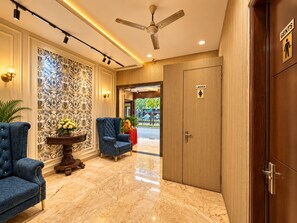 A lobby with a blue armchair, a decorative wall, a ceiling fan, and a door with a ladies' sign. - Hotel Taj Heights (Agra)