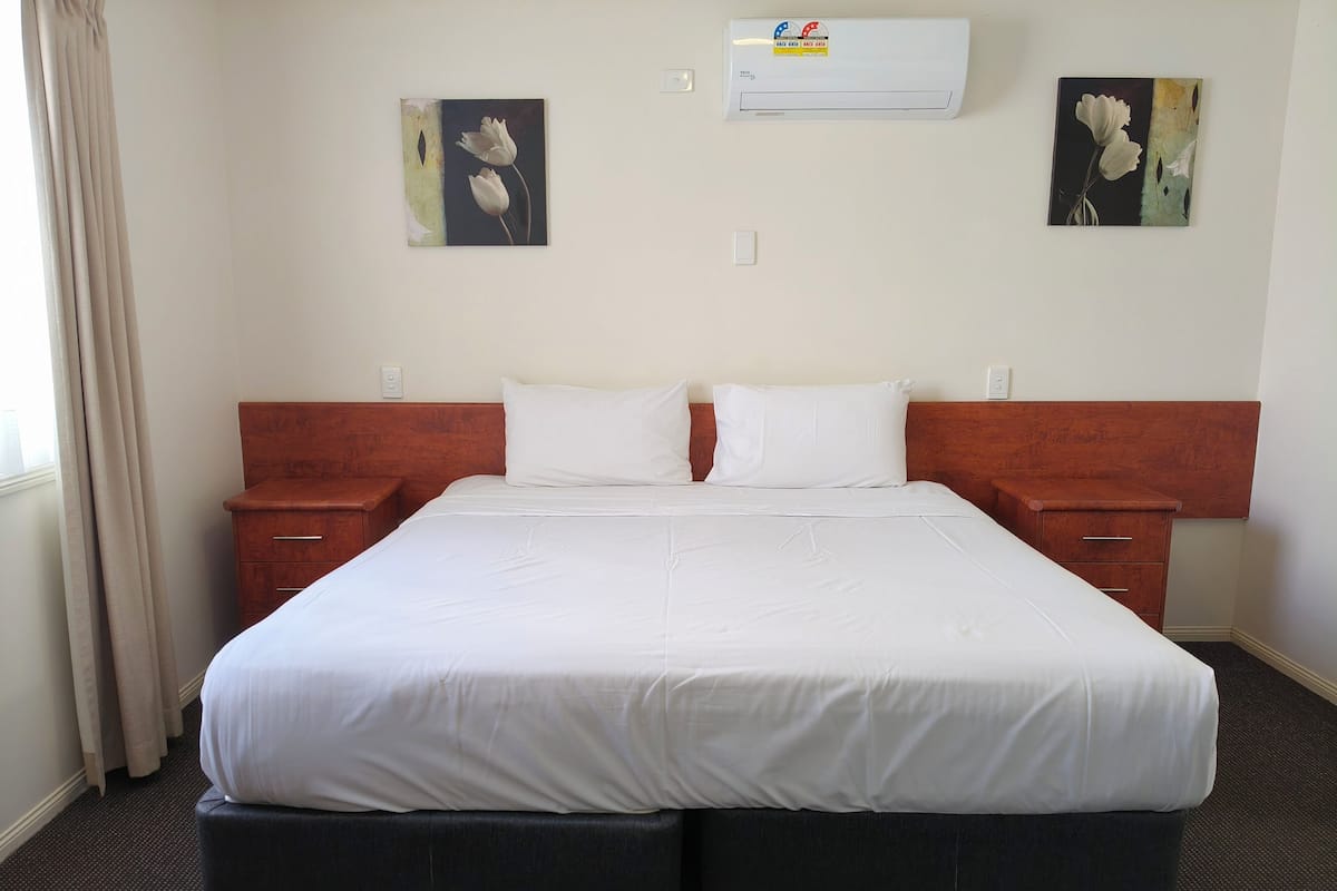 A hotel room with a large bed, two bedside tables, a wooden headboard, and two wall-mounted air conditioning units.