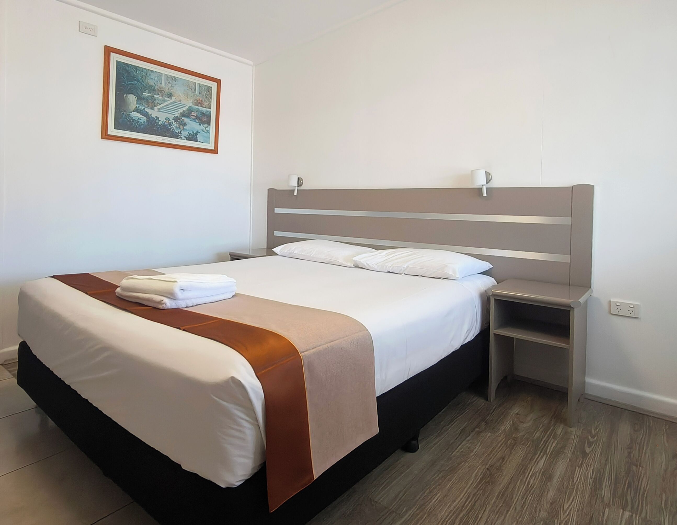 A neatly made bed with a brown accent, a bedside table, a wall-mounted lamp, and a framed picture above the bed.