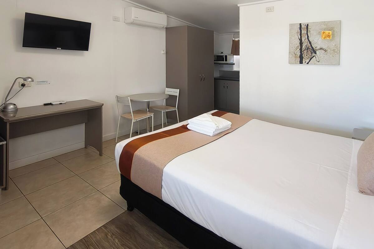 A hotel room with a bed, desk, chair, TV, and artwork on the wall.
